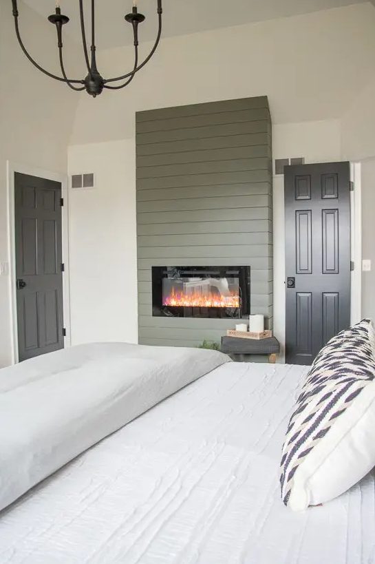 a beautiful farmhouse bedroom with a built-in fireplace with green planked wood covering, a bed with neutral bedding and a vintage metal chandelier
