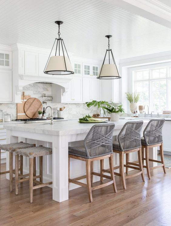 a beautiful modern kitchen with white shaker style cabinets, a large kitchen island with woven chairs and stools, pendant lamps