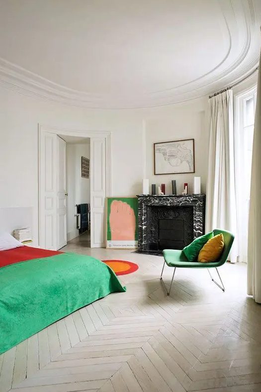 a bold bedroom with a fireplace clad with black marble, a bed with colorful bedding, a bold green chair and an artwork, a ceiling with molding