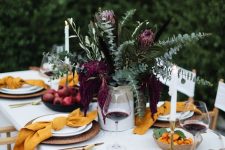 a bright modern Thanksgiving table with greenery and deep purple blooms, yellow napkins, woven chargers and gold cutlery rocks