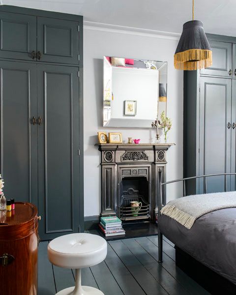 a chic bedroom with graphite grey wardrobes, a metal bed with grey bedding, a pendant lamp, a fireplace and a vanity with a stool