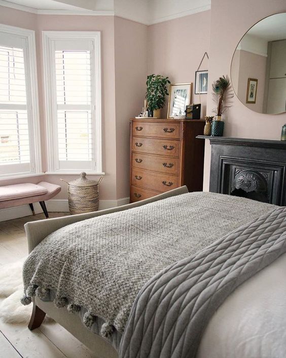 a chic bedroom with pink walls, a metal fireplace, a bed with grey bedding, a blush bench and a stained dresser