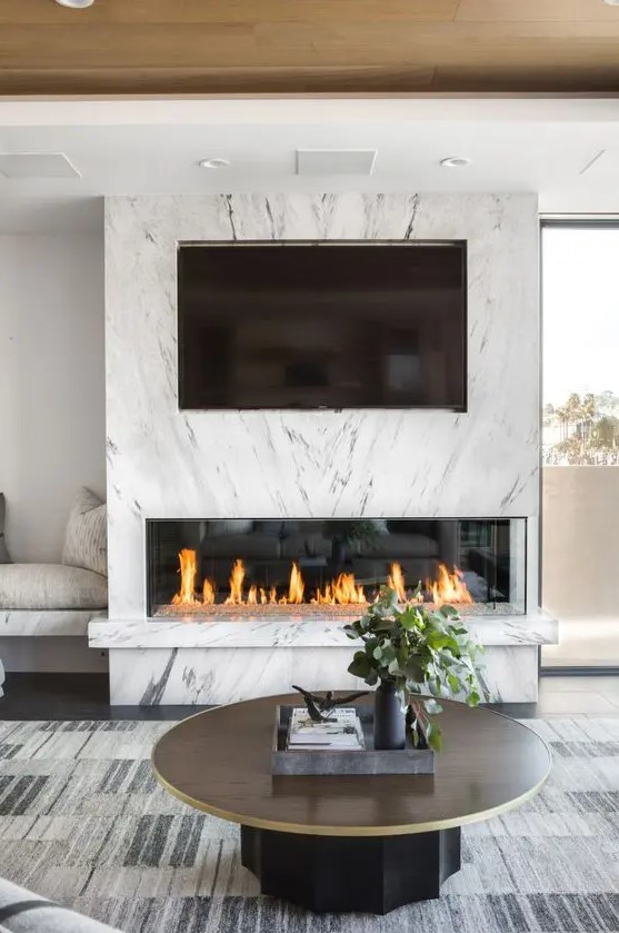 a chic modern space with a fireplace clad with white marble and a TV, a built-in bench, a round table and a statement rug
