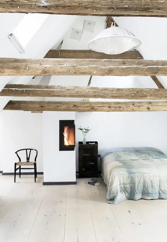 a cozy light-filled bedroom with wooden beams on the ceiling and a warming up hearth by the bed