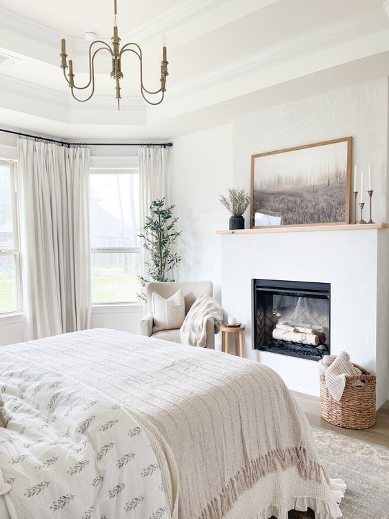 a creamy bedroom with a fireplace, a bed with neutral bedding, a creamy chair with a blanket, a basket and a chandelier