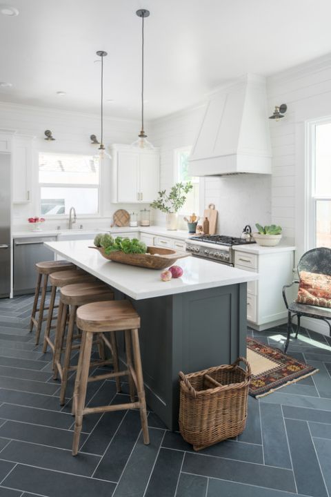 a farmhouse kitchen with white cabinets and a large hood, pendant lamps, a graphite grey kitchen island and wooden stools