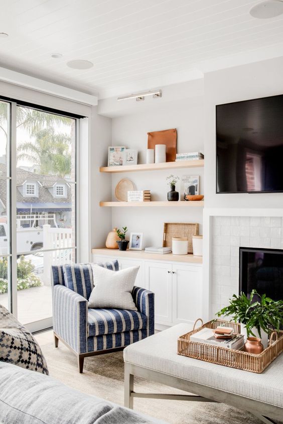 a farmhouse space wiht a white brick clad fireplace, built-in shelves and cabinets, a striped chair and a large ottoman and greenery