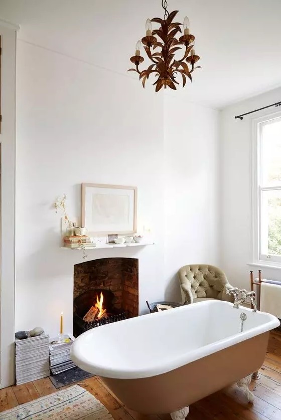 a gorgeous vintage bathroom in neutrals, with a fireplace clad with brick, a rust-colored bathtub, a chic chandelier and stacks of books