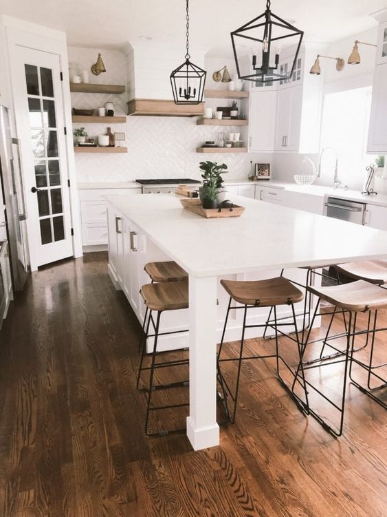 a large airy white kitchen with shaker style cabinets, stained shelves and a hood, a large kitchen island with a storage cabinet and an eating space