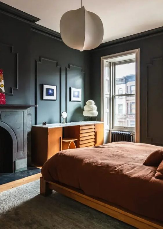 a lovely moody bedroom with black trim walls, a non-working fireplace, a stained bed with rust bedding, a stained desk