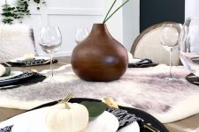 a modern Thanksgiving tablescape with a faux fur runner, black woven placemats, white porcelain and pumpkins, a brown vase with greenery