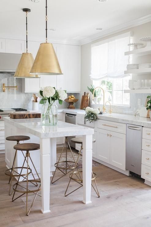 a modern and airy kitchen with white shaker style cabinets, a small kitchen island that doubles as a table, brass pendant lamps