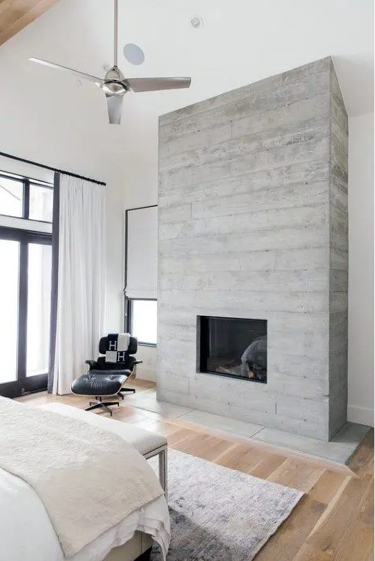 a modern bedroom in neutrals, with much natural light and a concrete fireplace that gives the space a cool look