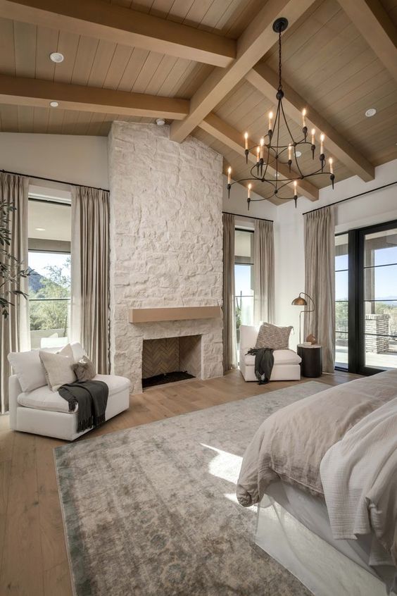 a modern chalet bedroom with a stone fireplace, a bed with neutral bedding, white chairs and neutral textiles