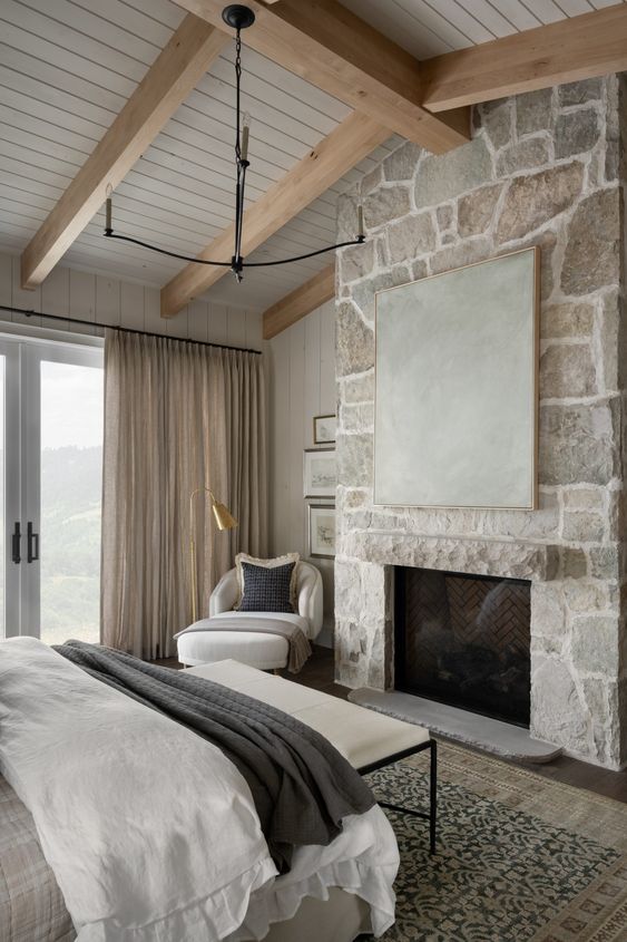 a modern farmhouse bedroom in neutrals, with a stone fireplace, a bed with neutral bedding, a white bench and a chair and a lovely view