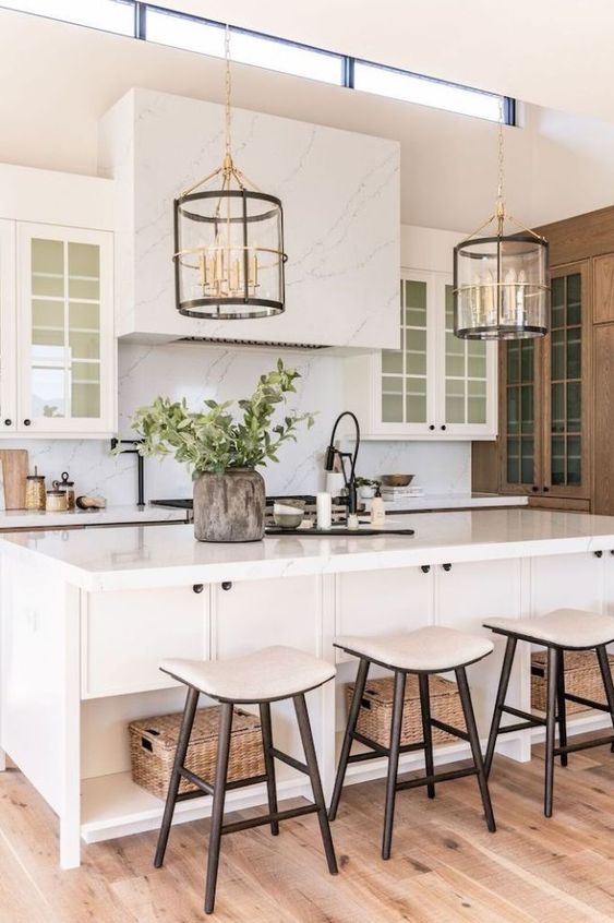 a pretty modern farmhouse kitchen done in creamy shades, with a white marble backsplash and a ahood, a large kitchen island and neutral stools