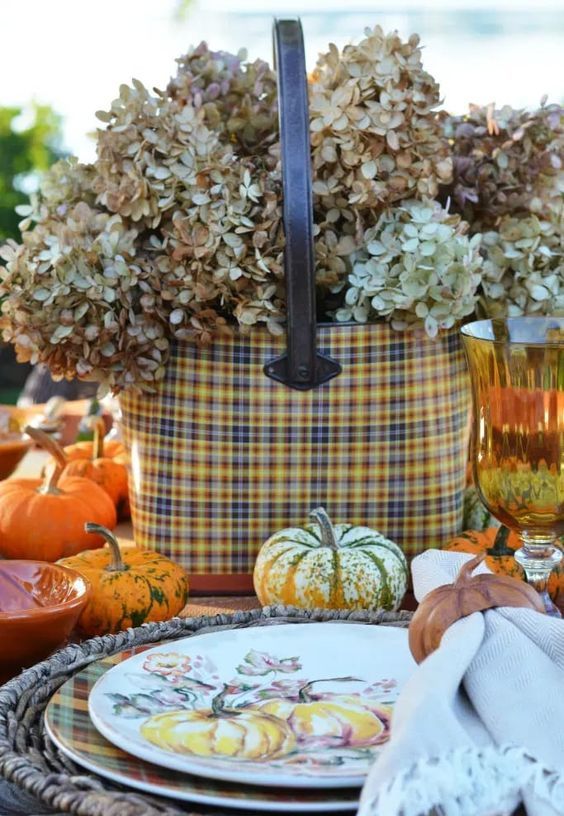 a pretty rustic Thanksgiving tablescape with woven placemats, plaid plates and a basket with dried hydrangeas, pumpkins and amber glasses