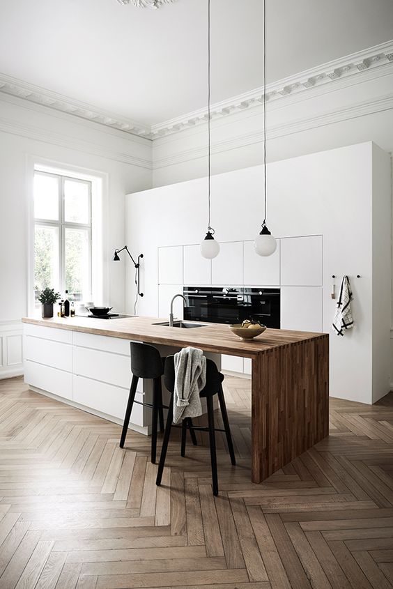 a refined minimalist kitchen with sleek white cabinets, a large kitchen island with a wooden waterfall countertop, black stools and pendant lamps