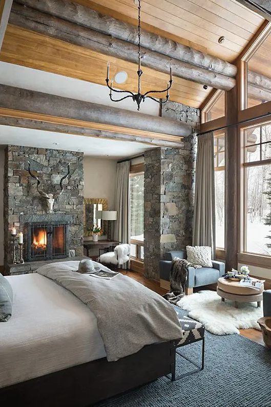 a rustic bedroom with wooden logs on the ceiling, a pillar clad with stone and a stone fireplace