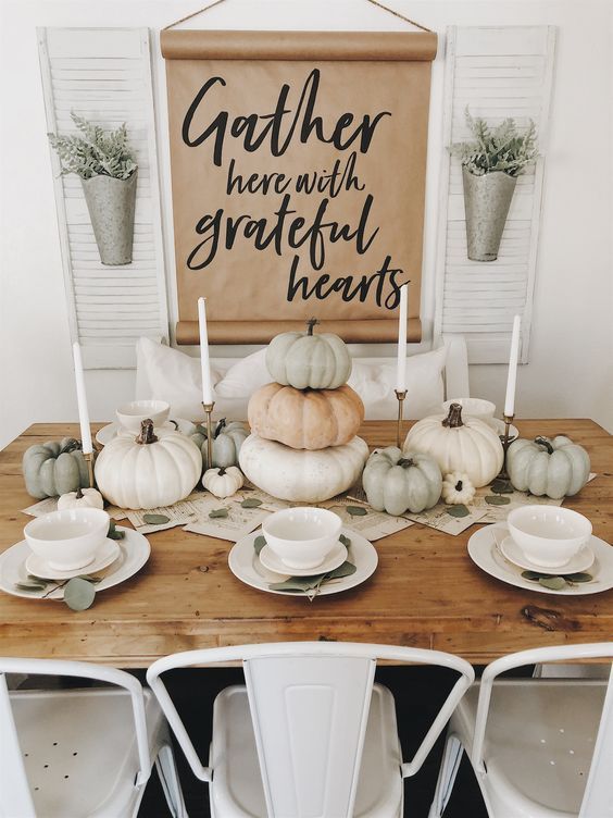 a simple and modern Thanksgiving table with white porcelain, neutral pumpkins stacked and some greenery on the table