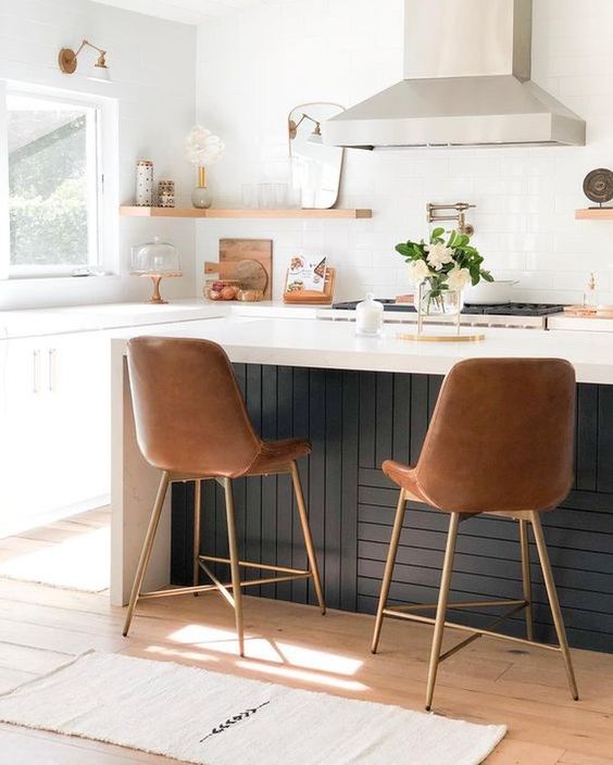 a small and pretty modern kitchen with white lower cabinets, open shelves, a black planked kitchen island and leather and brass stools