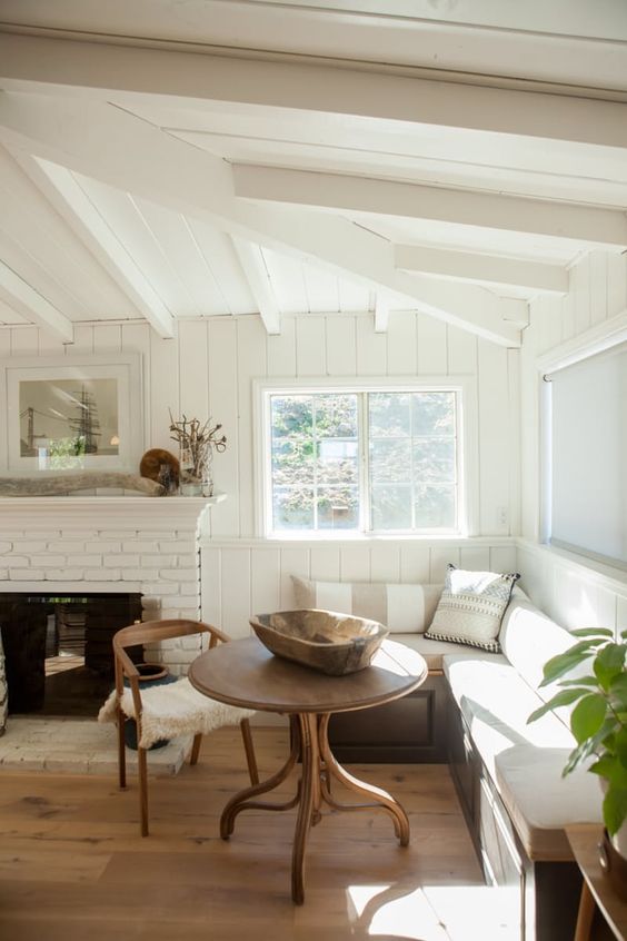 a white brick fireplace with a mantel, a corner banquette seating, a round table and a chair as a meal space by the fire