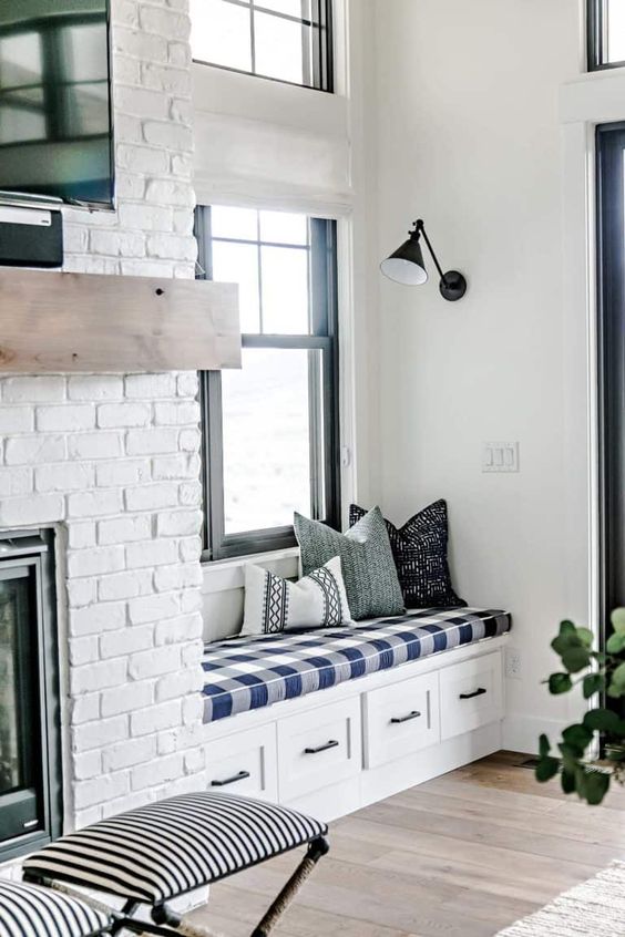a white farmhouse space with a brick fireplace, a wooden mantel, a built in bench with drawers, a plaid cushion and pillows