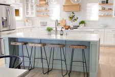 an airy kitchen with white shaker style cabinets, wooden shelves and a hood, a light blue kitchen island with wooden stools for eating