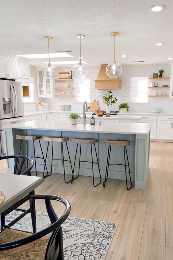 an airy kitchen with white shaker style cabinets, wooden shelves and a hood, a light blue kitchen island with wooden stools for eating