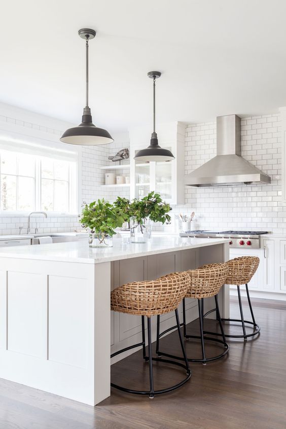 an airy white kitchen with a large kitchen island, black metal pendant lamps, woven stools on black metal bases and greenery
