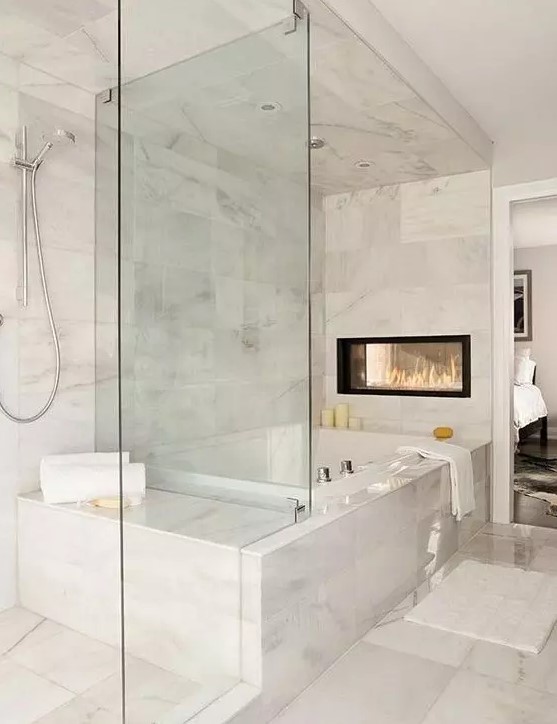 an exquisite white marble bathroom with a tub, a glass-enclosed shower space, a double-sided fireplace and some neutral textiles