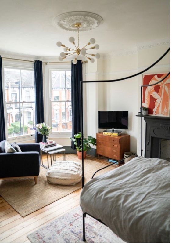 an eye-catchy bedroom with a bay window, a fireplace, a black chair and tables, a TV zone and a metal frame bed