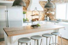 an industrial kitchen with white cabinetry, a brick wall, a planked kitchen island and metal stools plus metal pendant lamps