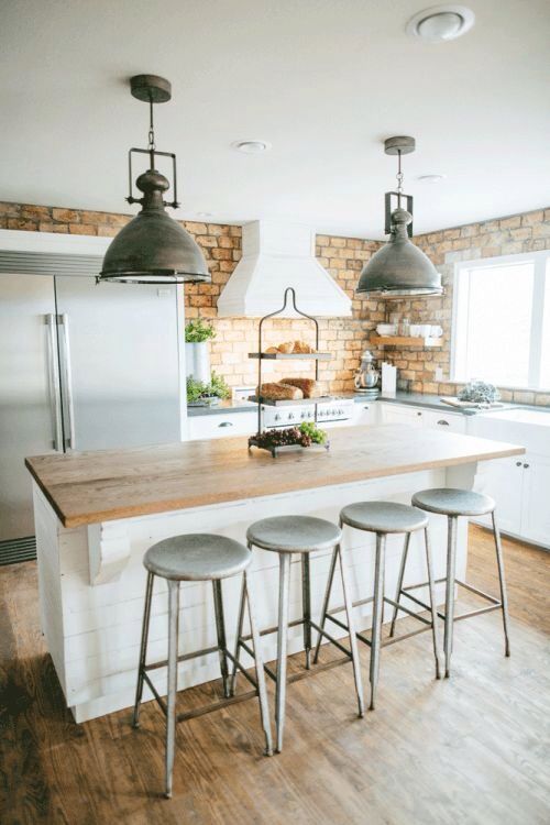 an industrial kitchen with white cabinetry, a brick wall, a planked kitchen island and metal stools plus metal pendant lamps