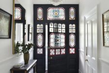 a Victorian townhouse entryway with a black and white tiled floor, a black and gold console table, a mirror and a black door and sidelights with stained glass