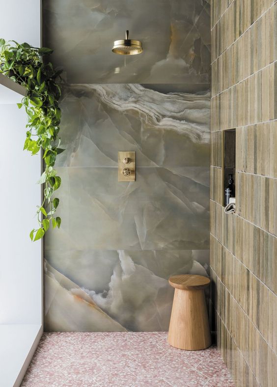 a chic contemporary shower space with green onyx tiles and a tan narrow tile wall, a wooden stool and brass fixtures