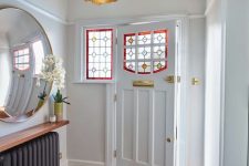 a chic vintage-inspired entryway with a black radiator, a shelf and a large mirror, a printed rug and a door with stained glass plus gold touches
