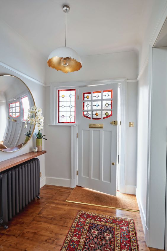 a chic vintage-inspired entryway with a black radiator, a shelf and a large mirror, a printed rug and a door with stained glass plus gold touches