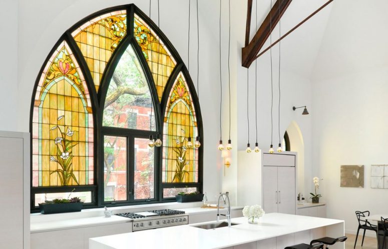 a contemporary to minimalist kitchen done in white, with wooden beams and bulbs hanging and a unique Gothic-style window with stained glass