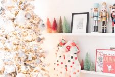 a flocked Christmas tree with white and silver ornaments, colorful pompom stockings and colorful tinsel Christmas trees on the shelf