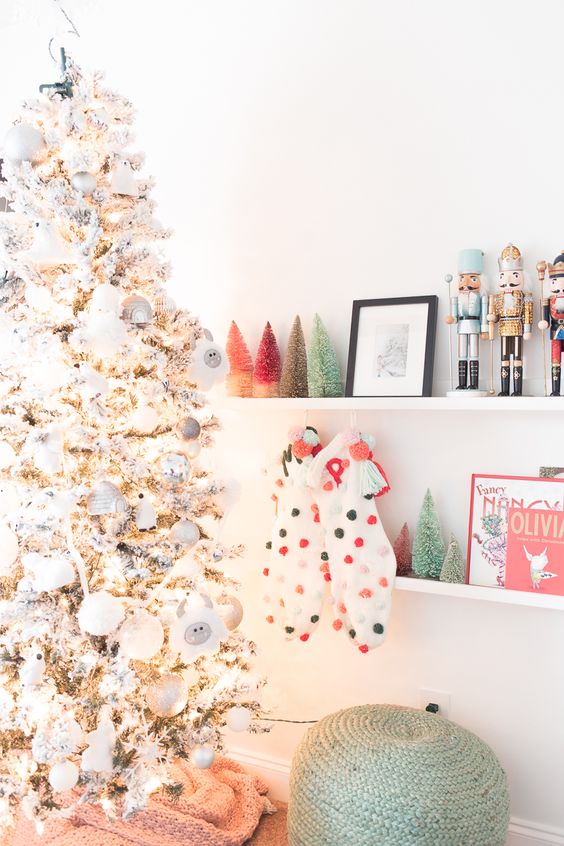 a flocked Christmas tree with white and silver ornaments, colorful pompom stockings and colorful tinsel Christmas trees on the shelf