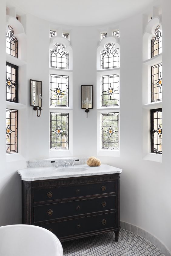 a neutral bathroom with small window with stained glass, a black vintage vanity, mirrors with lights and a tub is very chic