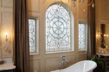 a vintage bathroom with patterned tan walls, a vintage bow window with stained glass, a clawfoot bathtub, potted blooms and a chandelier