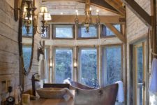 a chic chalet bathroom with wooden walls and beams, a glazed wall, a metal tub and chic chandeliers and lamps