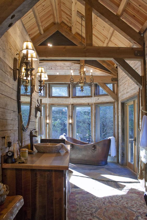 a chic chalet bathroom with wooden walls and beams, a glazed wall, a metal tub and chic chandeliers and lamps