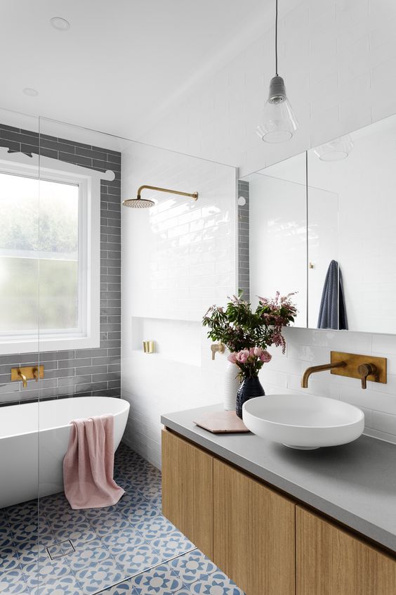 a chic contemporary bathroom with grey, white and blue patterned tiles, a floating vanity and a round sink, brass fixtures and a window with frosted glass