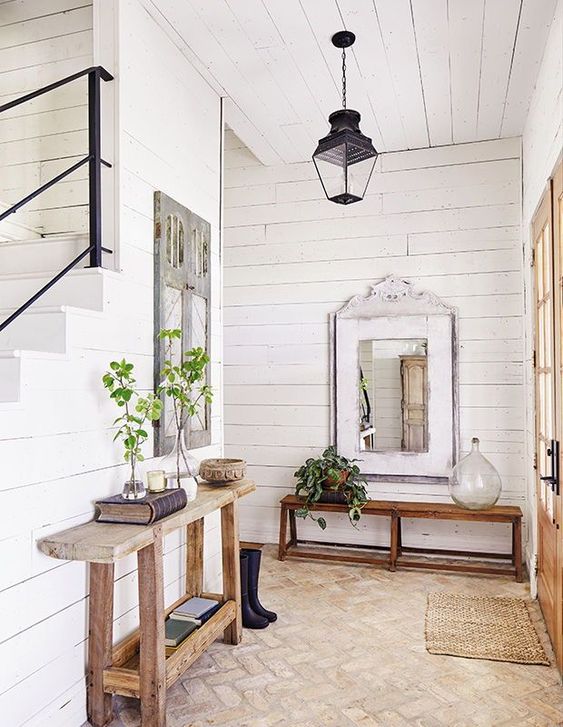 a farmhouse entryway with a console and a bench, a lamp, a jute rug, some greenery in vases and mirrors