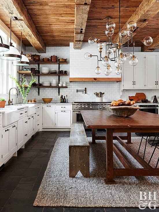 a refined chic kitchen in white, with stylish cabinetry, a wooden table and benches, a wooden ceiling with beams and elegant chandeliers