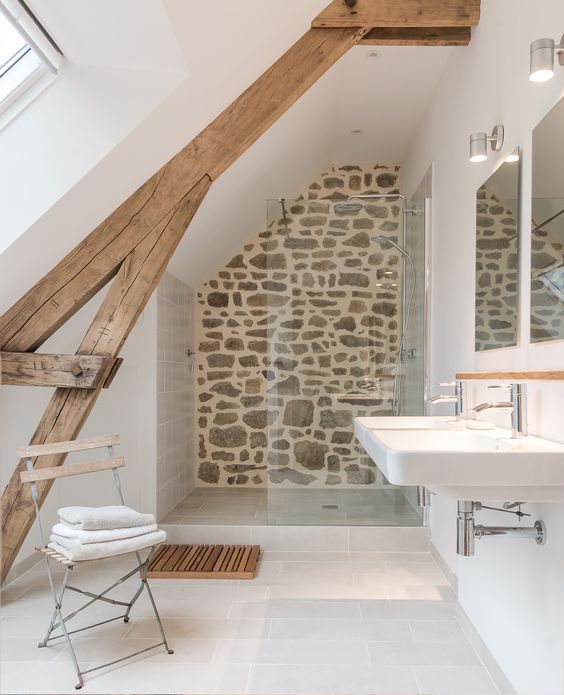 a small neutral bathroom with wooden beams, a stone wall, a wall mounted sink and a wooden chair plus a window
