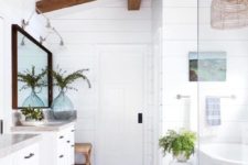 a white coastal bathroom with rich stained wooden beams, two vanities, a bathtub and a shower space plus greenery